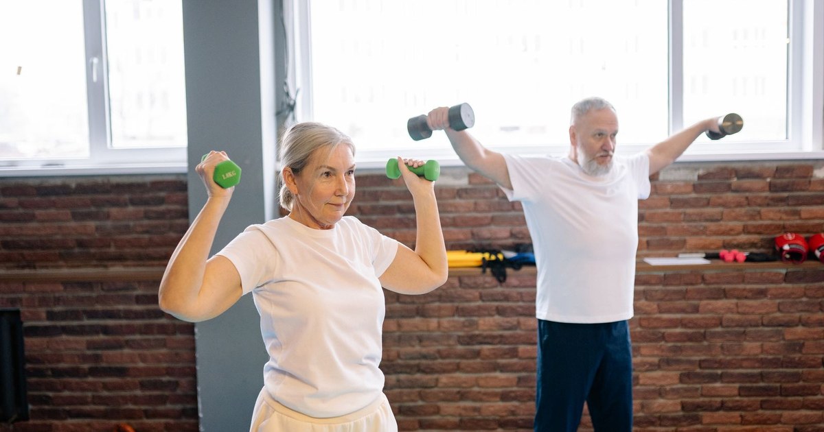 An older woman and an older man in white T-shirts raising small dumbbells in a bright gym with a brick wall