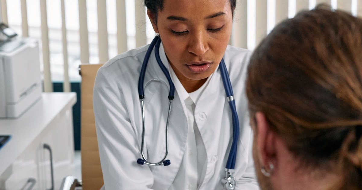 A doctor in a white coat with a stethoscope speaking with a patient during a clinic consultation