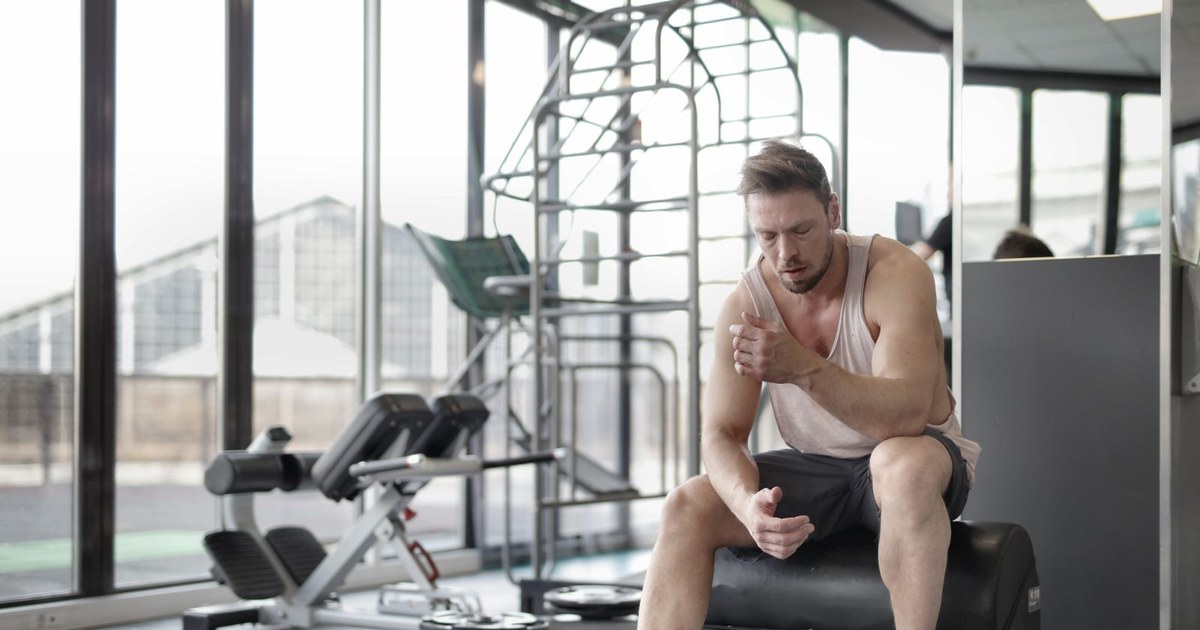 A muscular male athlete in workout clothes sitting on a bench and resting after a weightlifting session in a modern gym
