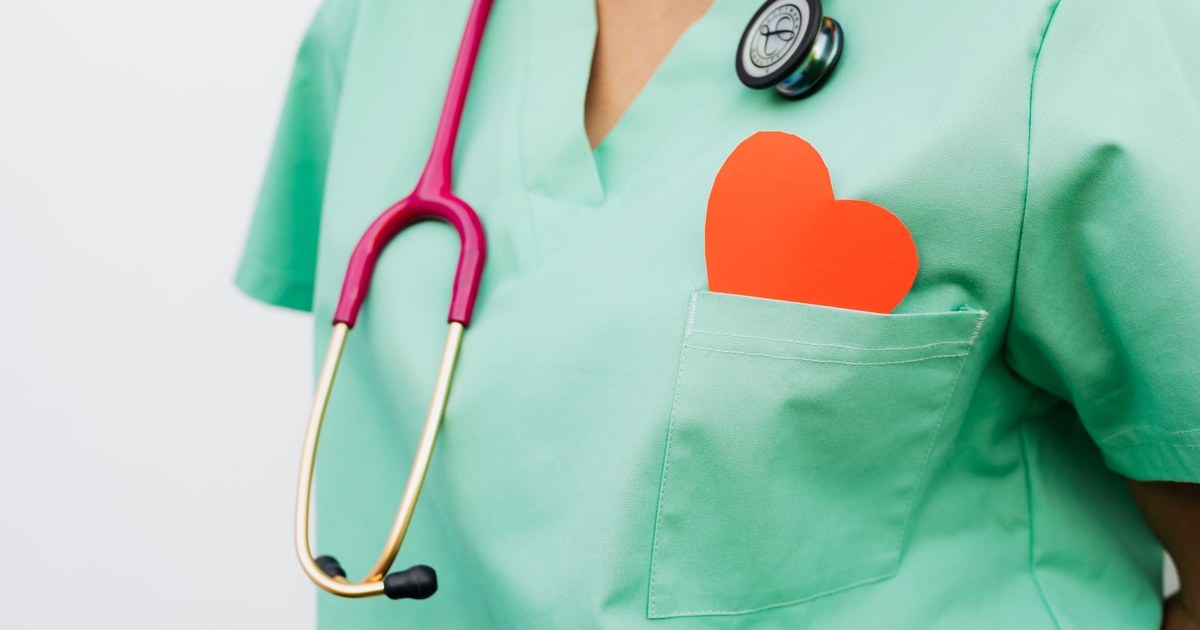 A healthcare worker in green scrubs wearing a stethoscope, with a small red paper heart in the chest pocket