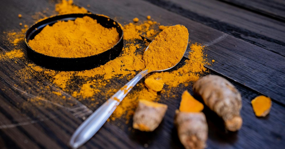 Golden turmeric powder in a bowl with a spoon and fresh turmeric root on dark wood