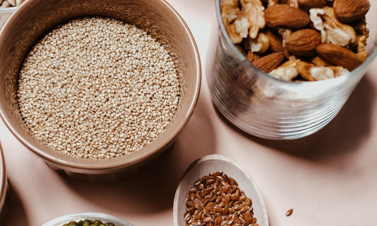 Bowls of magnesium-rich foods including pumpkin seeds, quinoa, mung beans, almonds, walnuts, and flax seeds on a soft pink surface